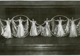 Teenage girls in shimmering dresses dance on stage with two large rings as props.