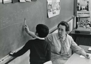 Historical photo of an older teacher, sitting in a chair, working with a young boy to solve the math math problem written on the blackboard behind them.
