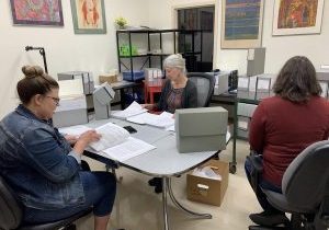 Three women sit in an office meeting room sorting papers.