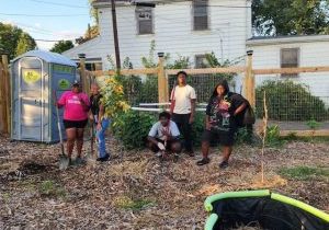 One woman in a pink shirt stands with four young people holding garden equipment standing next to a tree they planted in their garden.