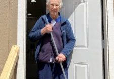 An older woman with white hair stands in the doorway of a shed holding a broom looking at the camera.