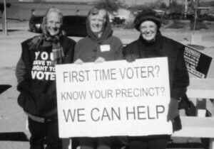 A black and white photo of three older women dressed in very warm clothes standing outside smiling holding a sign that says "First time voter? Know your precinct? We can help"