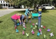 Three women work together to decorate a large patch of grass with handmade paper windmills. The field also has a bright teal wooden wheelbarrow with assorted flowers and a hand-painted pole surrounded by the paper windmills.