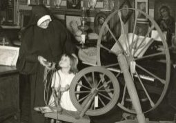 Black and white photo of a nun in habit looking down at a young girl. They stand next to a spinning wheel in a room surrounded by Christian art and artifacts.