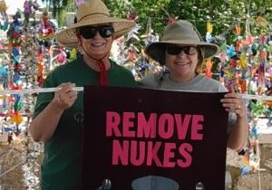 Two women in hats and sunglassed hold a "Remove Nukes" banner in front of strands of paper cranes.